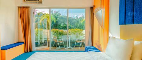 A hotel room with a large bed, a balcony with a glass door, and a view of greenery outside.