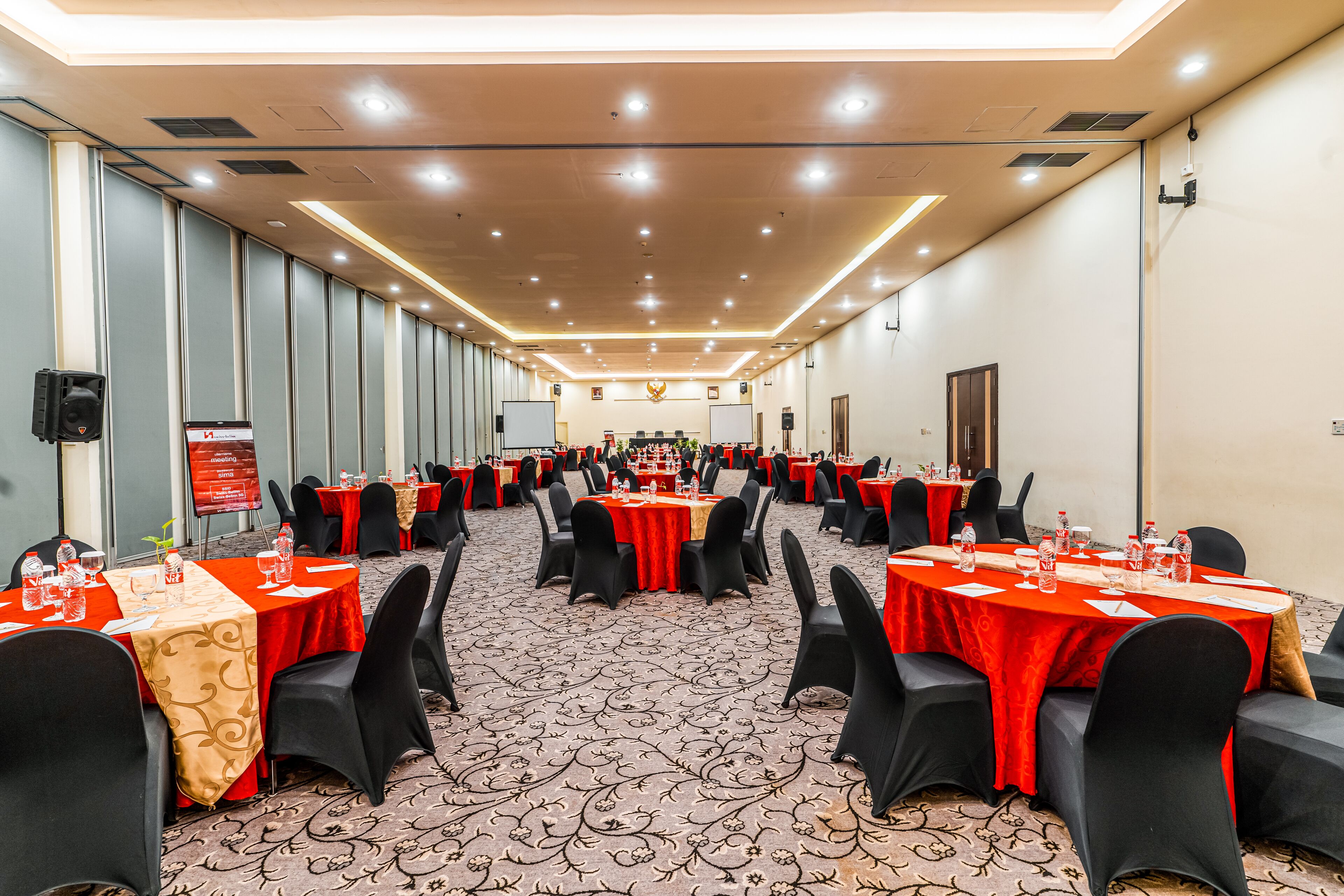 Un salón de conferencias con varias mesas, sillas negras y manteles rojos.
