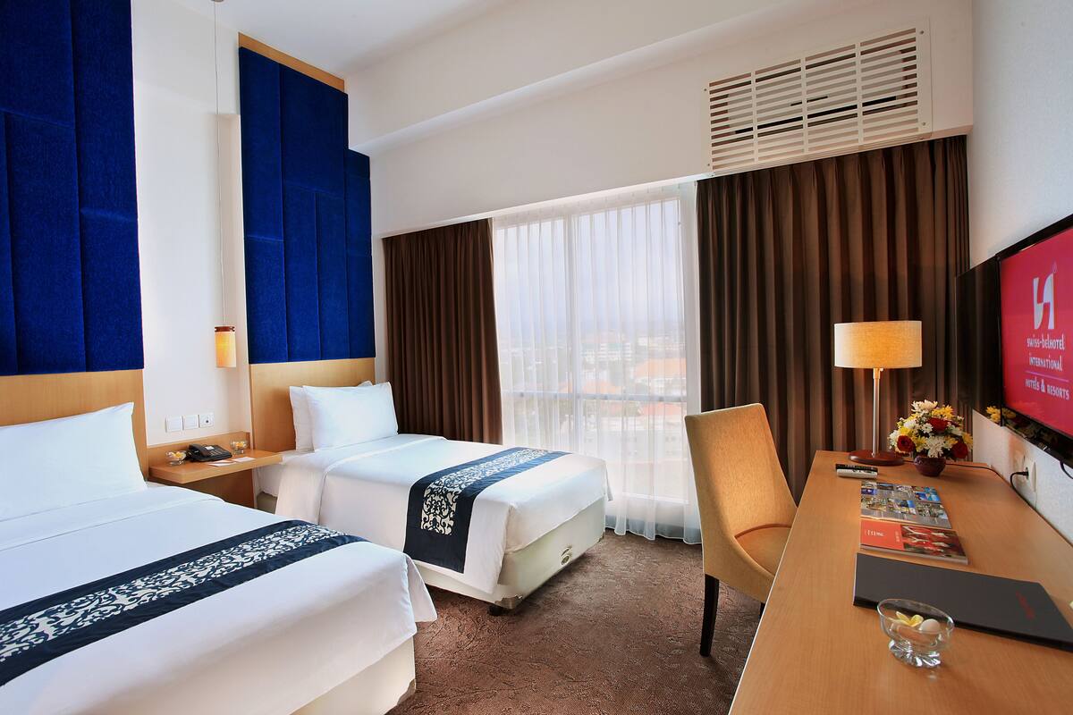 Une chambre d’hôtel avec deux lits, un bureau avec une chaise, une télévision et une fenêtre avec des rideaux.