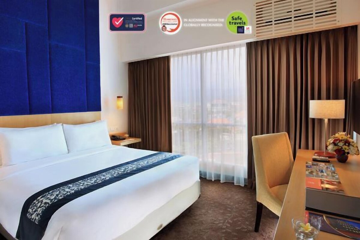 Una habitación de hotel con cama, escritorio, silla, lámpara y ventana con cortinas.