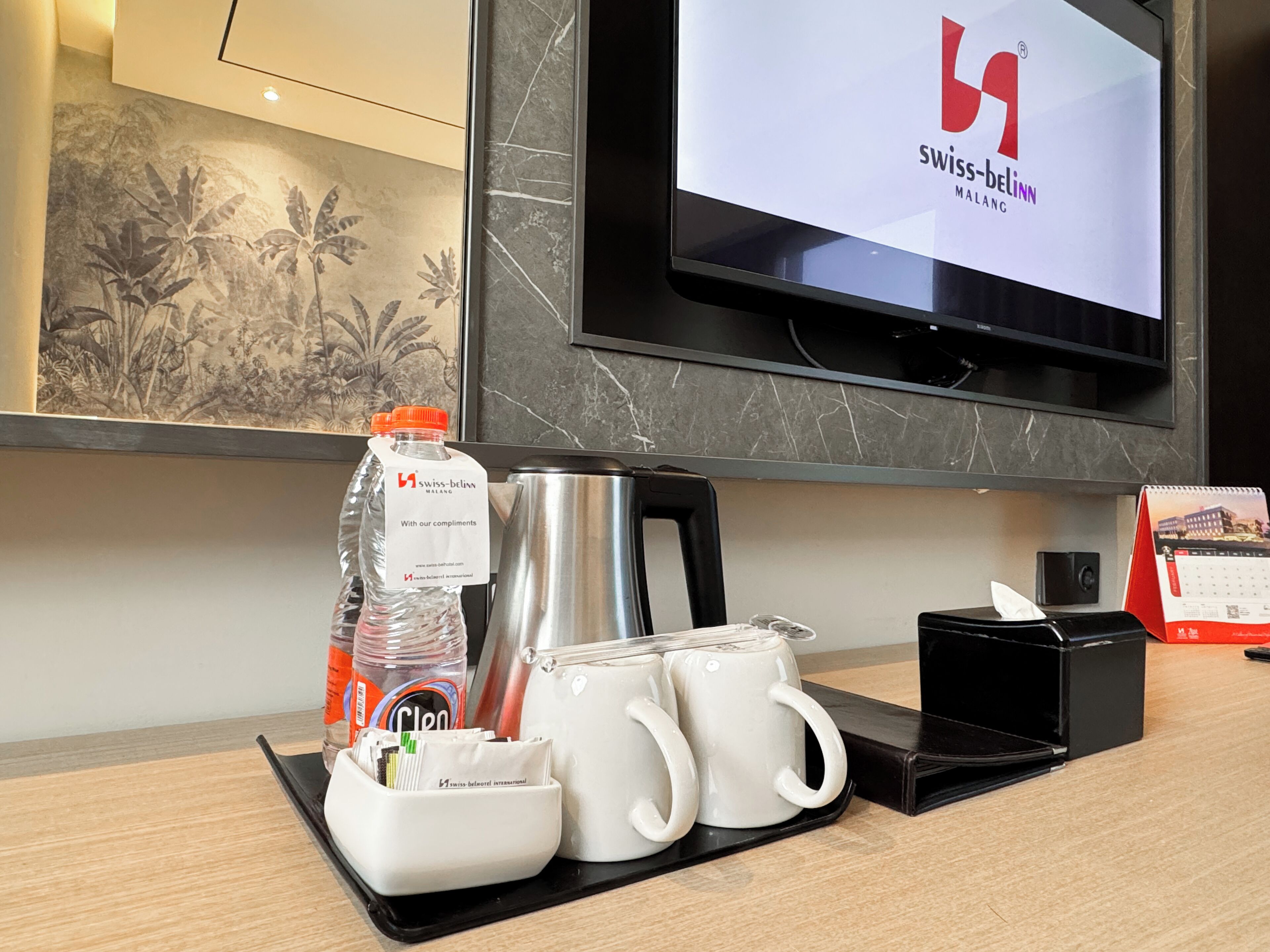 Hotelkamer met een televisie waarop het Swiss-Belinn logo staat, een koffiezetapparaat, twee mokken, een waterfles, een pot koekjes en een kalender.