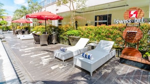 An outdoor lounge area with white wicker lounge chairs and red umbrellas, a wooden deck, and a building with a 'SWISS BELLE' sign. - Swiss-Belinn Malang (Malang)