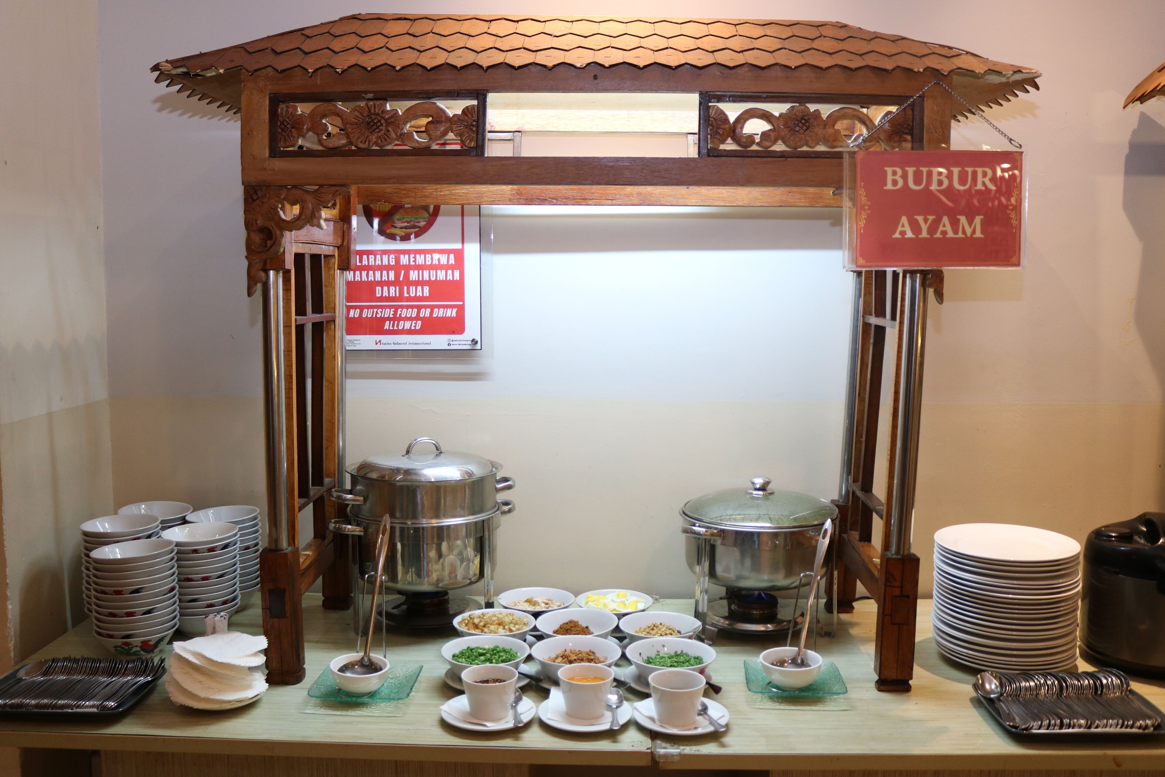 Montaje de bufé con varias estaciones de comida, incluyendo un cartel que indica 'BUBUR AYAM' (arroz con huevo).