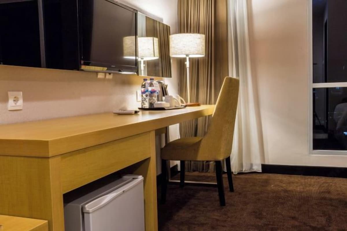Habitación de hotel con escritorio de madera, una silla, una lámpara, un refrigerador pequeño y un ventanal con cortinas.