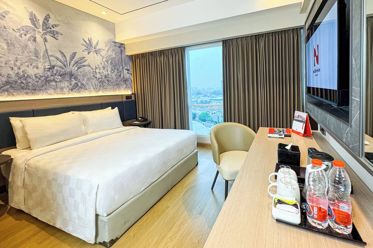 Une chambre d’hôtel comprenant un lit, un bureau avec une chaise, une télévision et une vue sur la ville grâce à de grandes fenêtres.