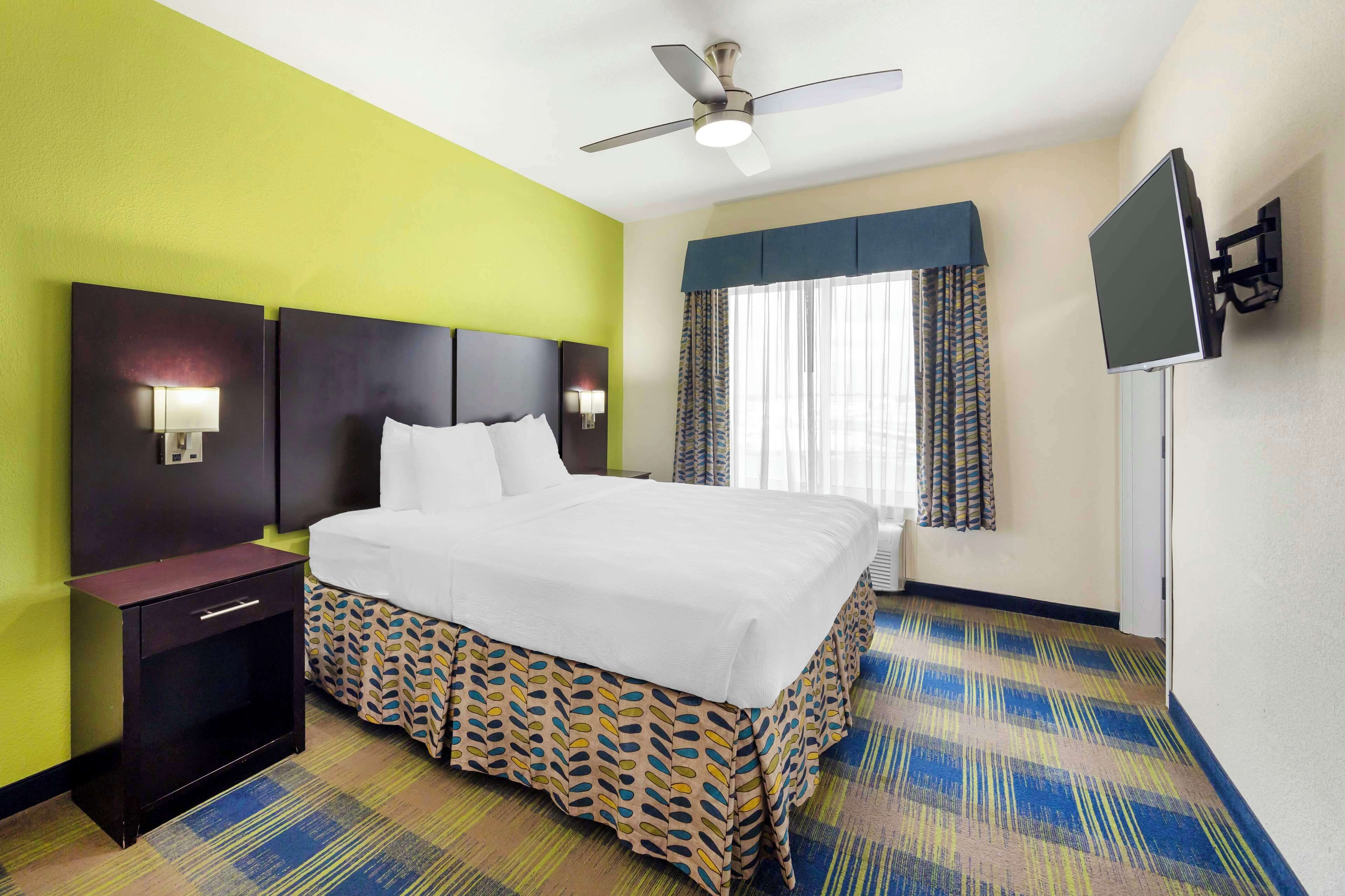 Habitación de hotel con cama, mesita de noche, televisor montado en la pared, ventilador de techo y alfombra estampada.