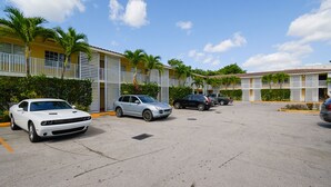 A parking lot with several cars, including a white sports car and a silver SUV, in front of a row of yellow buildings with balconies and palm trees. - Gables Cove Hotel (Coral Gables)