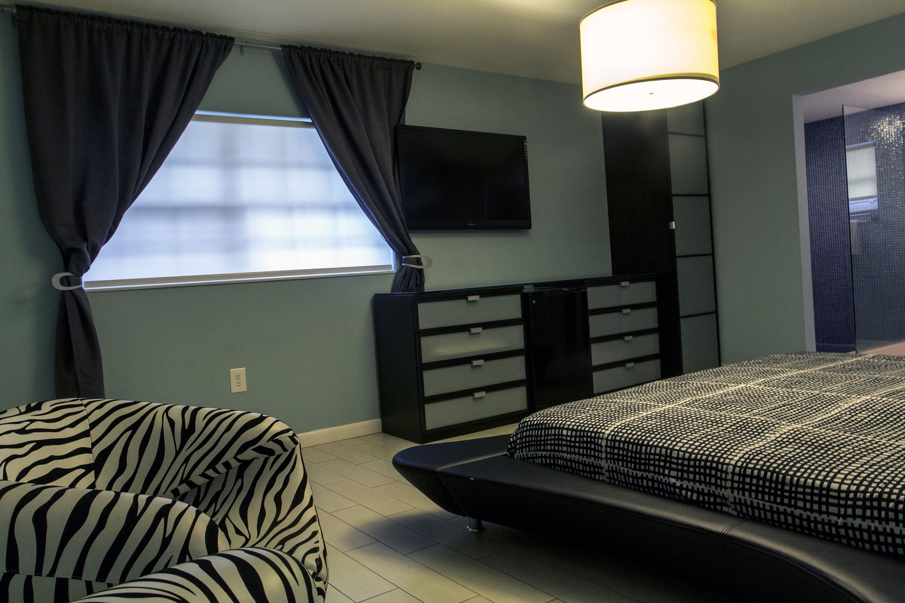 A bedroom with a bed, a chair, a TV, and a window with curtains.