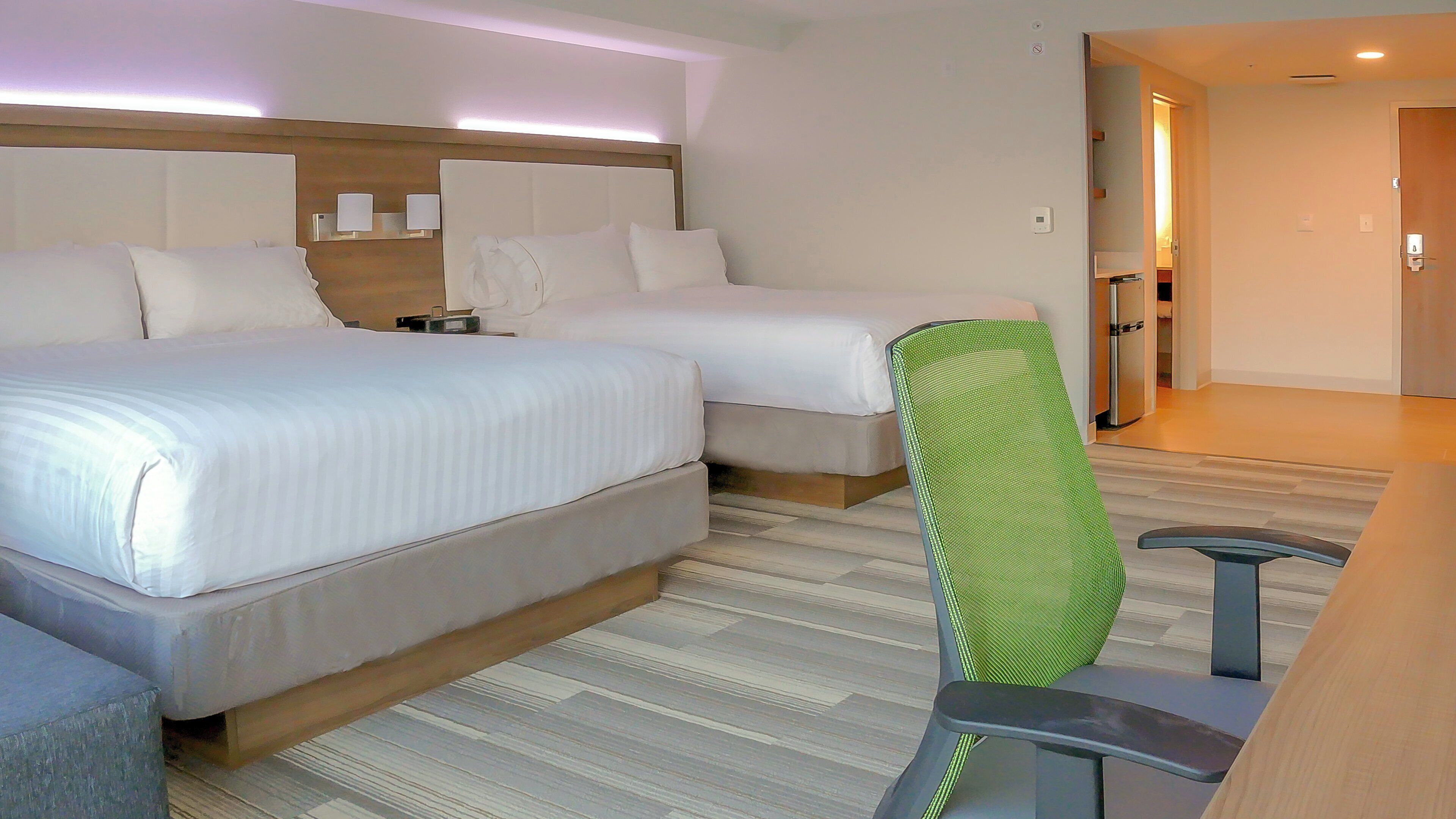 A hotel room with two beds, a desk, a chair, and a kitchenette.