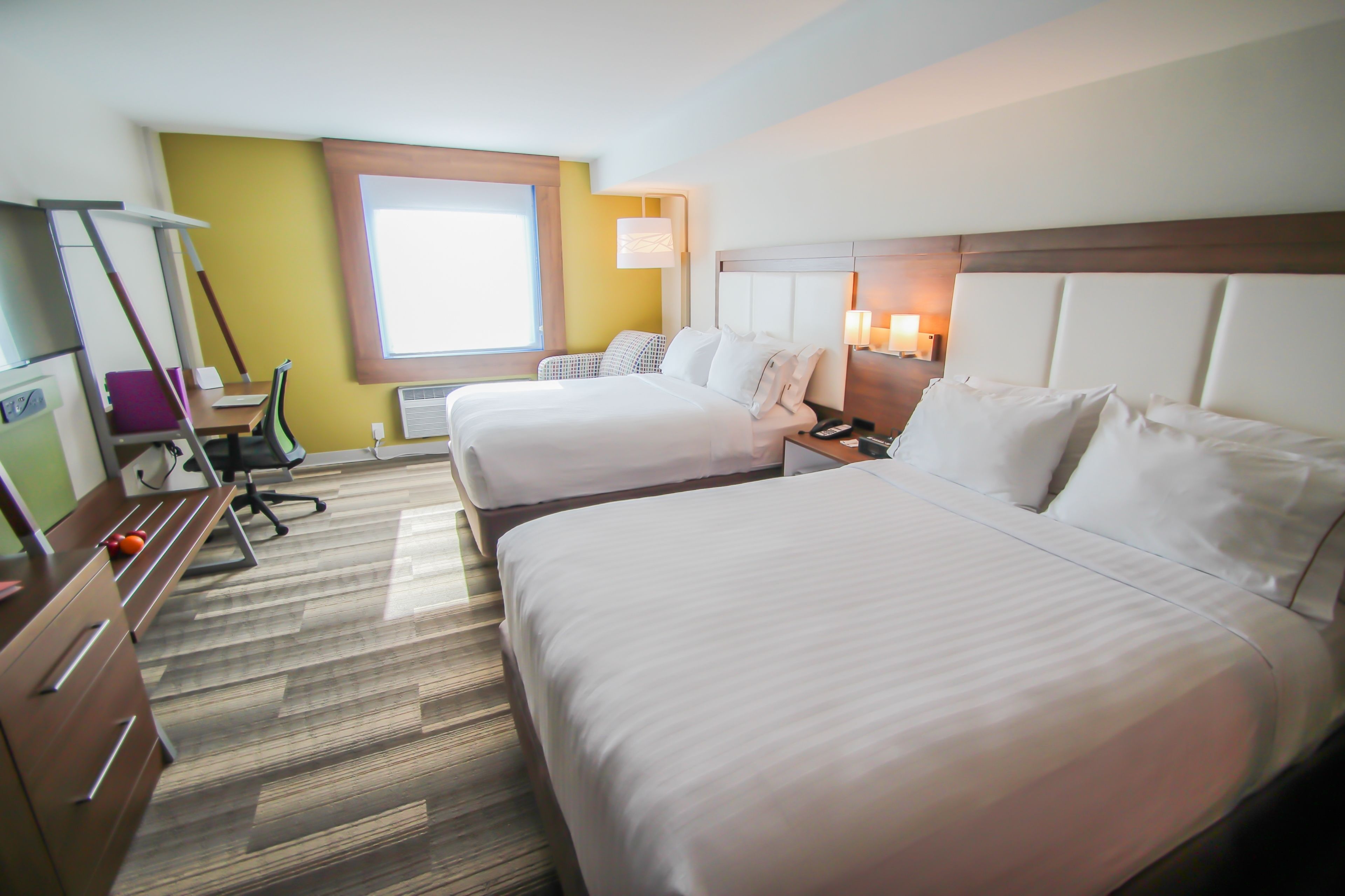 A hotel room with two beds, a desk, a chair, a window, and a wall-mounted lamp.