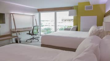 Habitación de hotel con dos camas, un escritorio, una silla y una ventana con vistas a edificios y árboles.