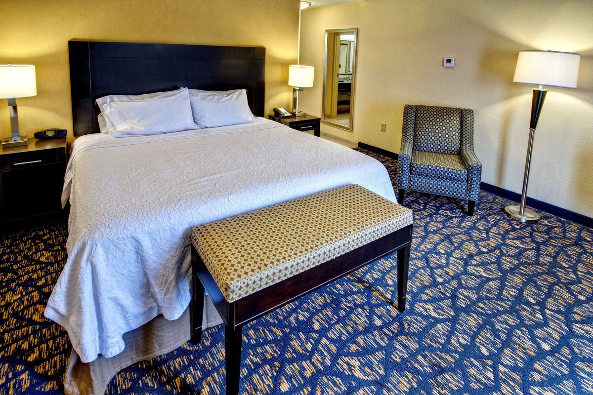 A hotel room with a large bed, a bedside table with a lamp, a chair, a bench, and a patterned carpet.
