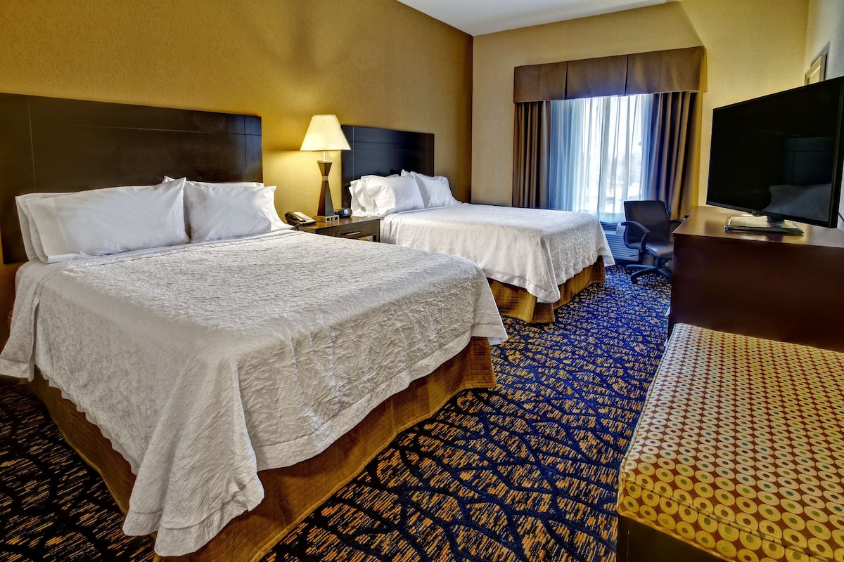 A hotel room with two beds, a desk, a chair, a TV, and a window with curtains.
