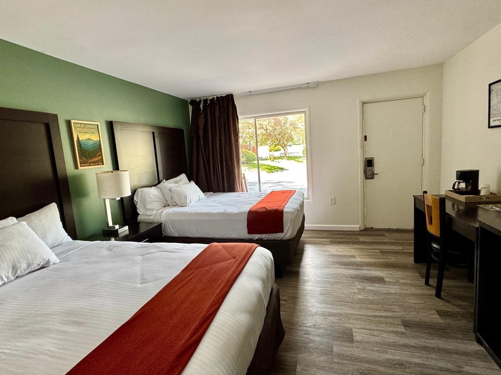 A hotel room with two beds, a desk, and a chair. There is a window with curtains, a door, and a wall-mounted lamp.