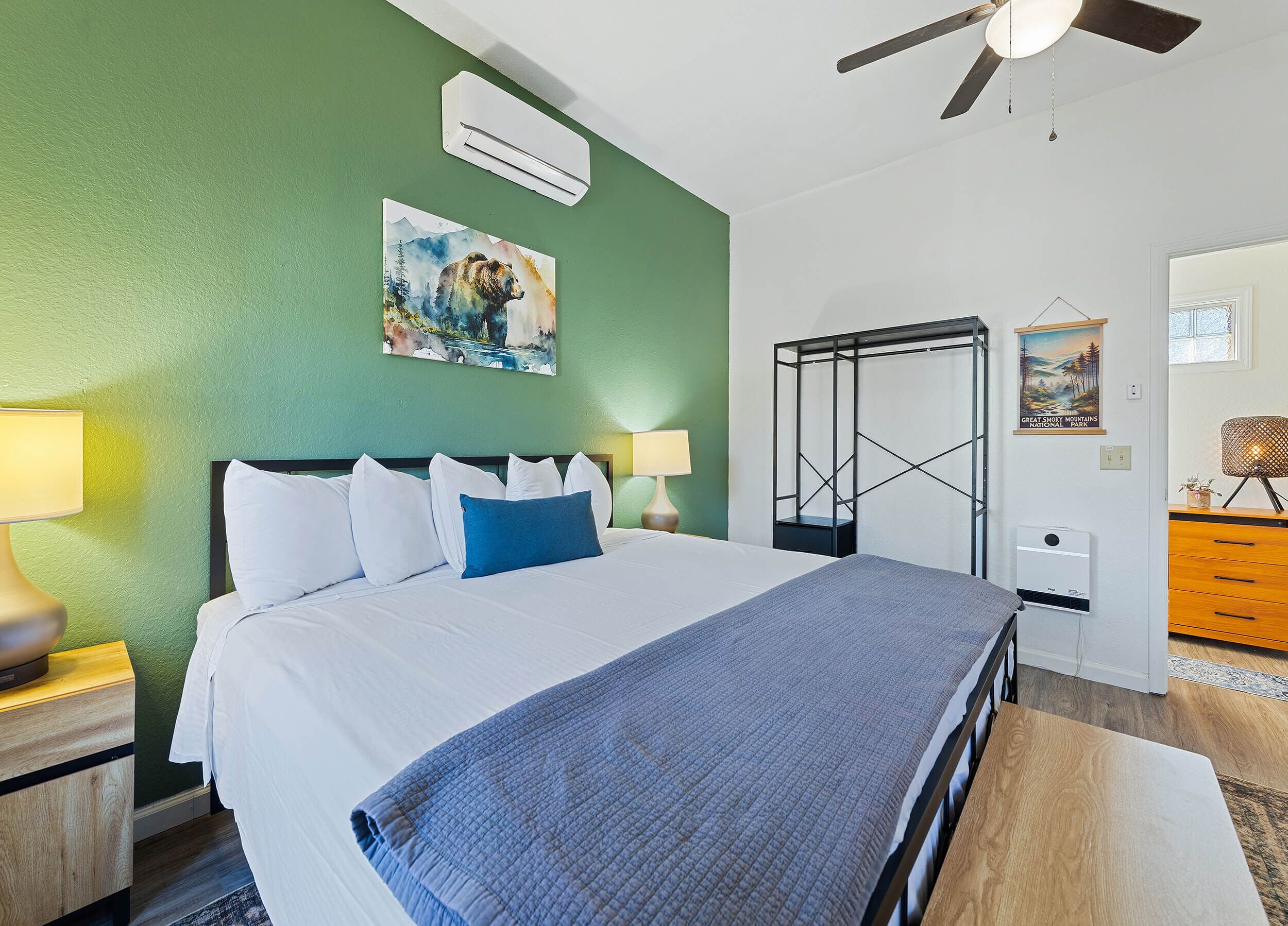 A hotel room with a bed, two bedside lamps, an air conditioner, a ceiling fan, a painting, a dresser, and a door.