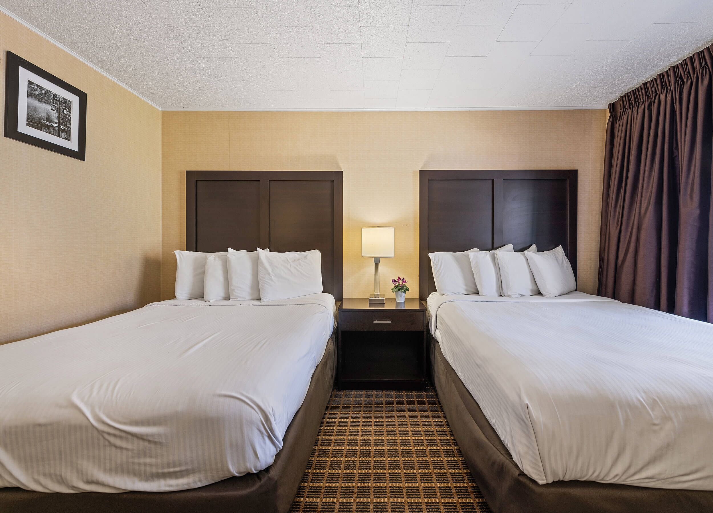 A hotel room with two beds, a nightstand, a lamp, and a picture on the wall.