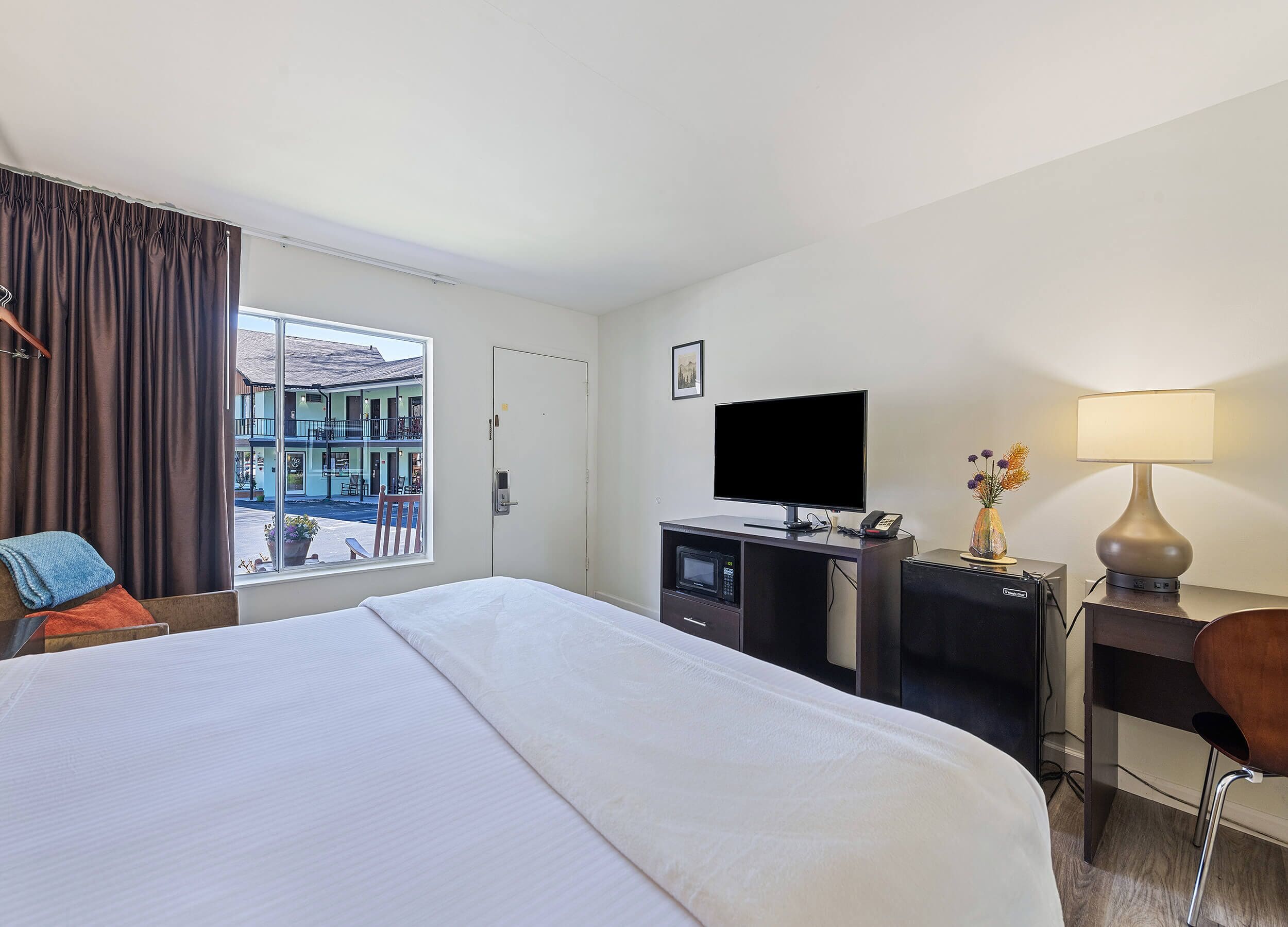 A hotel room with a bed, a desk with a computer, a TV, a lamp, and a view of the street.