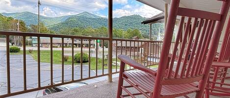 A wooden rocking chair on a covered balcony with a view of a parking lot and mountains in the background.