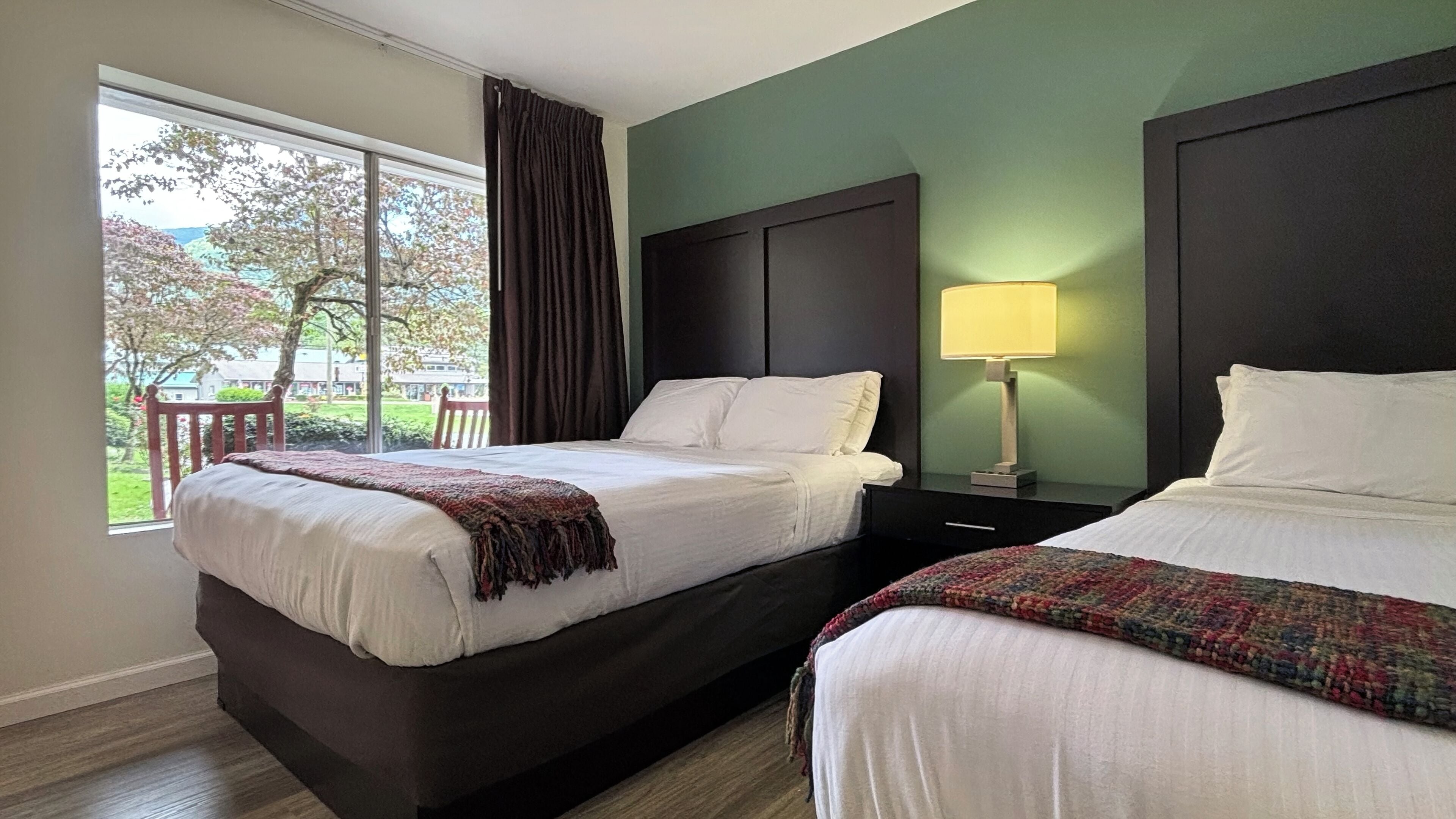 Habitación de hotel con dos camas, ventana que da a árboles, cabecera oscura y mesita de noche con lámpara.