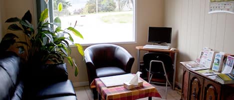 A living room with a black leather sofa, a wooden cabinet, a small table with a checkered tablecloth, a computer desk with a monitor, and a large window with a view of greenery outside.
