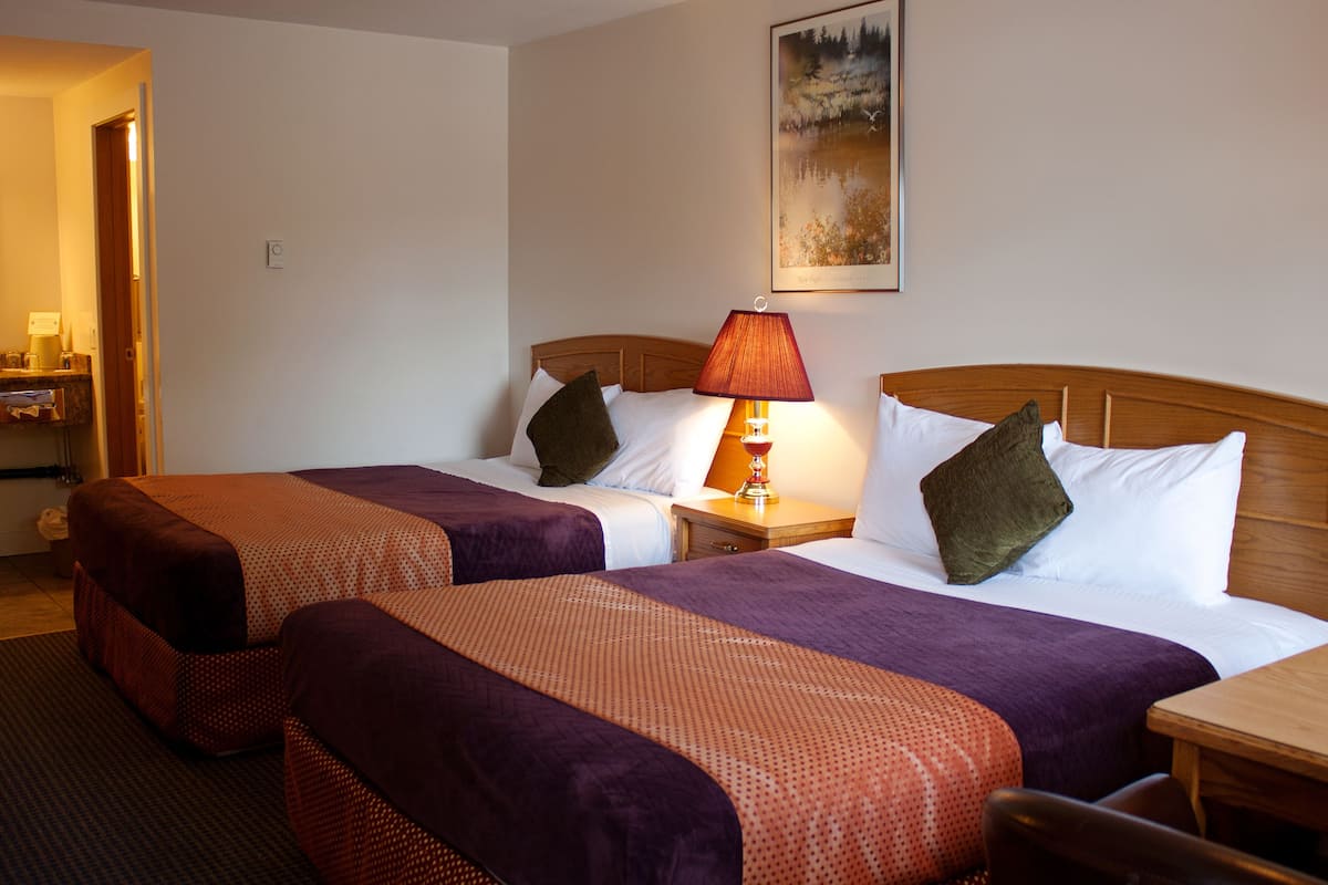 A hotel room with two beds, a nightstand with a lamp, a painting on the wall, and a wooden table.