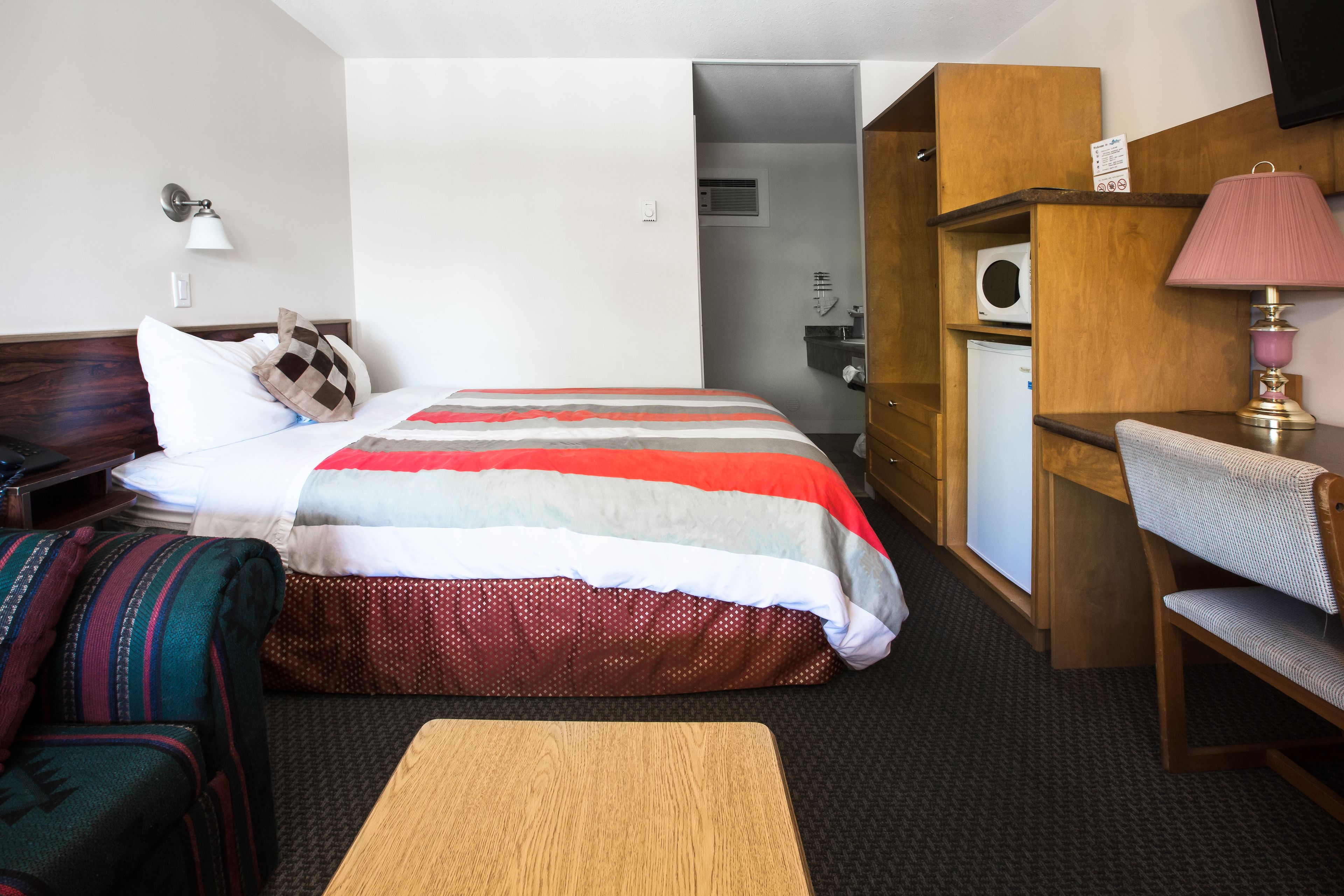 A hotel room with a bed, a desk, a chair, a lamp, and a small refrigerator.