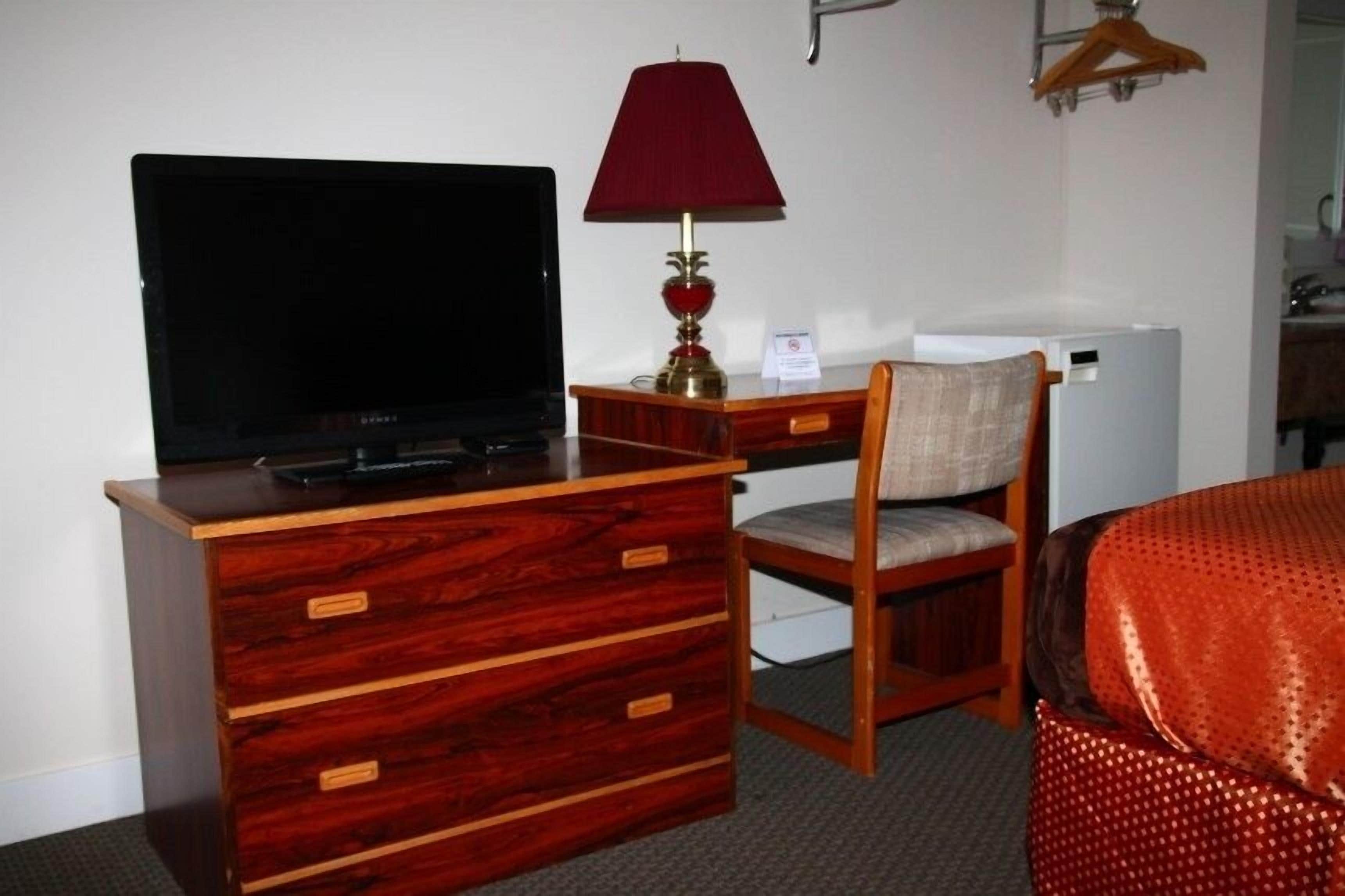 A hotel room with a wooden dresser, a television, a red lamp, a chair, and a bed with a red patterned cover.