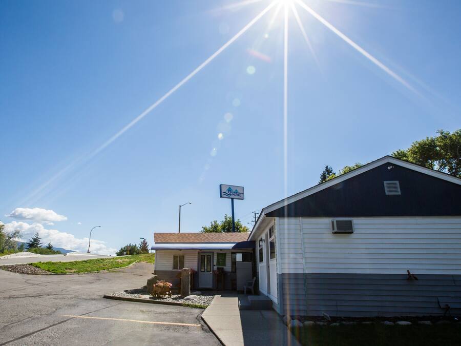 Un motel con aparcamiento, un pequeño edificio con un letrero y una casa grande con techo oscuro.