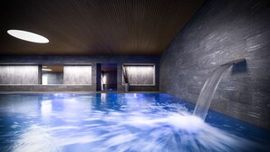 An indoor swimming pool with a waterfall feature, wooden ceiling, and large windows. - Valbella Resort (Vaz-Obervaz)