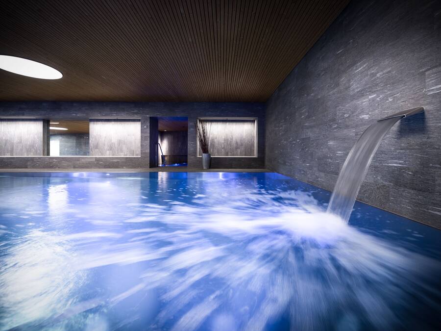 Una piscina interior con caída de agua, techo de madera y ventanas amplias.