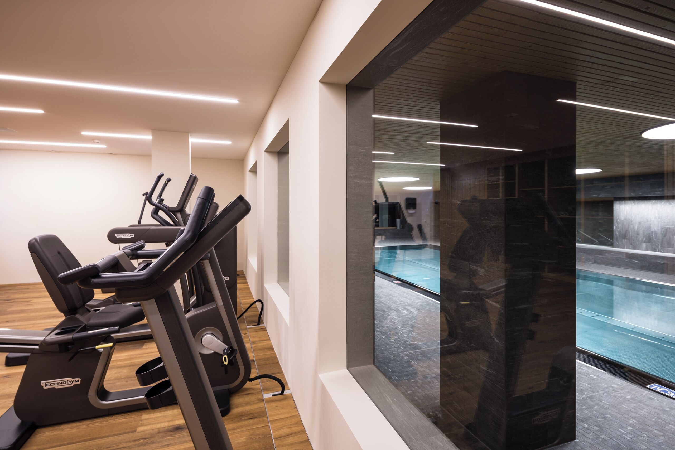 Un gimnasio moderno con máquinas de cardio, un ventanal con vistas a una piscina y un diseño limpio y minimalista.