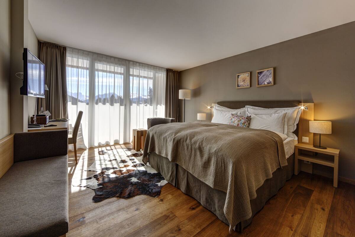 Ein modernes Hotelzimmer mit einem großen Bett, einem Schreibtisch mit Computer, einem Flachbildfernseher und Bergblick durch transparente Vorhänge.