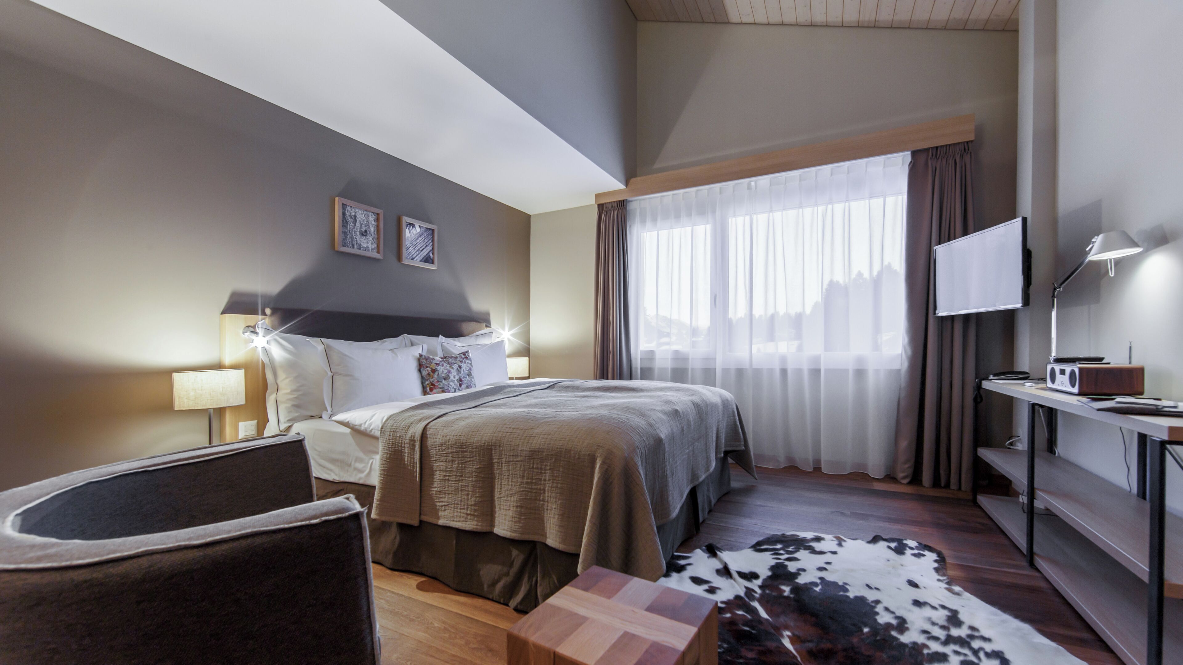 A hotel room with a bed, a desk with a lamp, a chair, a television, and a large window with curtains.