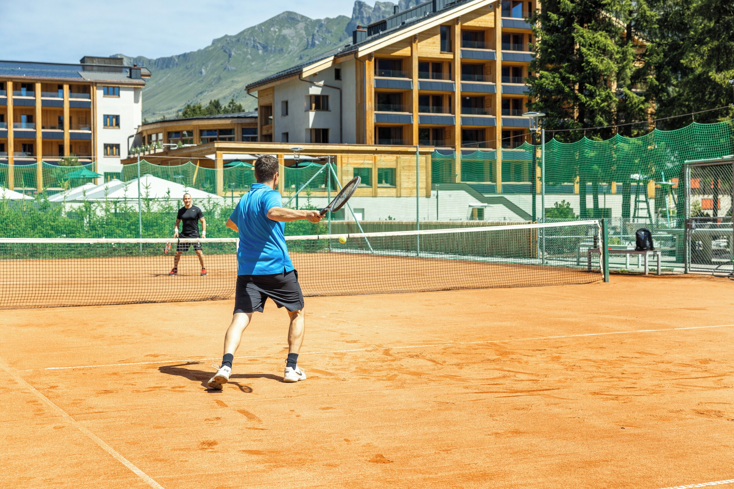Un campo da tennis con una rete, due giocatori e un edificio con balconi sullo sfondo.