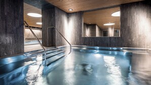 An indoor swimming pool with a wooden ceiling, dark walls, and a metal staircase leading into the pool. - Valbella Resort (Vaz-Obervaz)