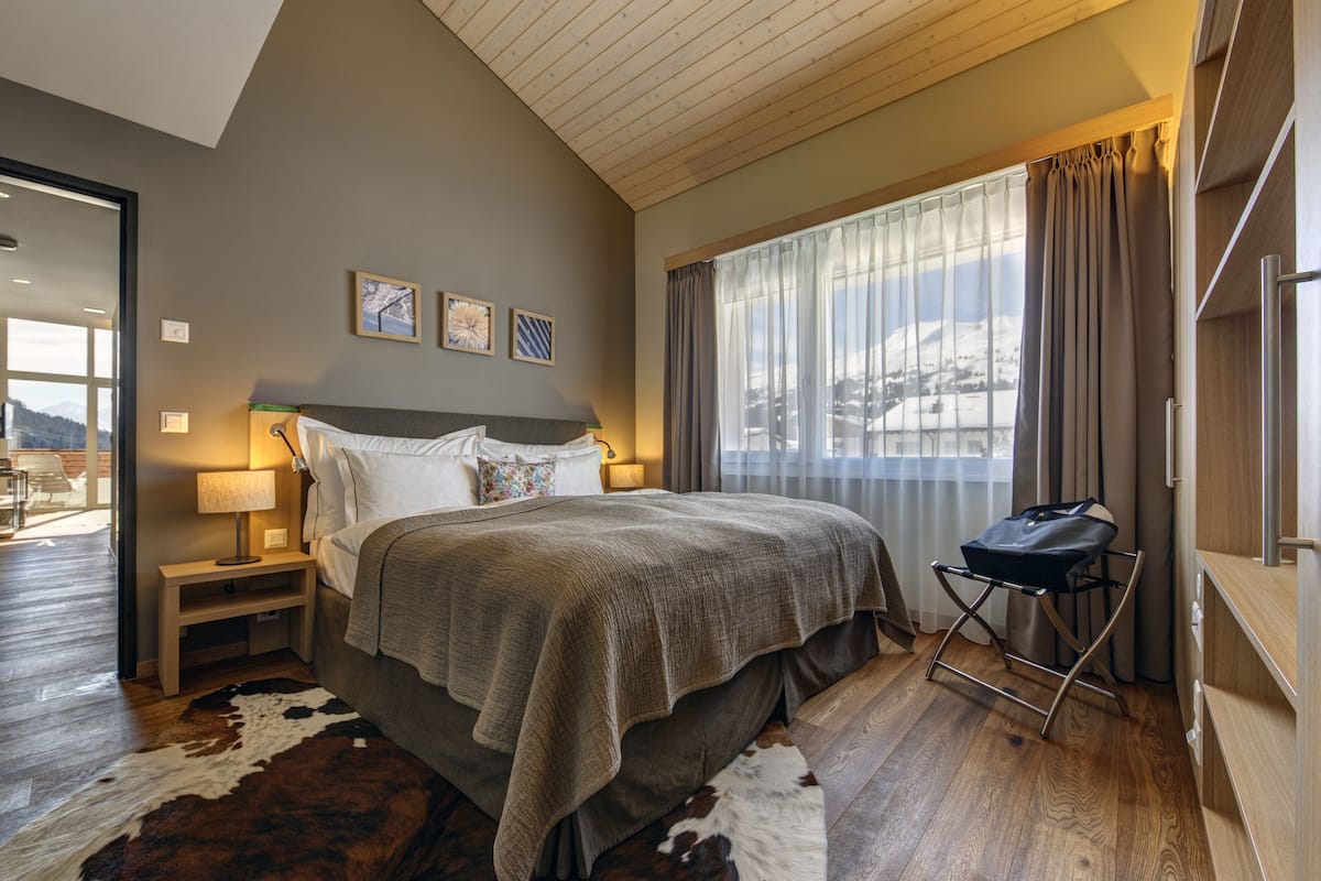 Ein Schlafzimmer mit einem großen Bett, einem Nachttisch, einer Lampe, einem Fenster mit Vorhängen und Blick auf eine verschneite Landschaft.