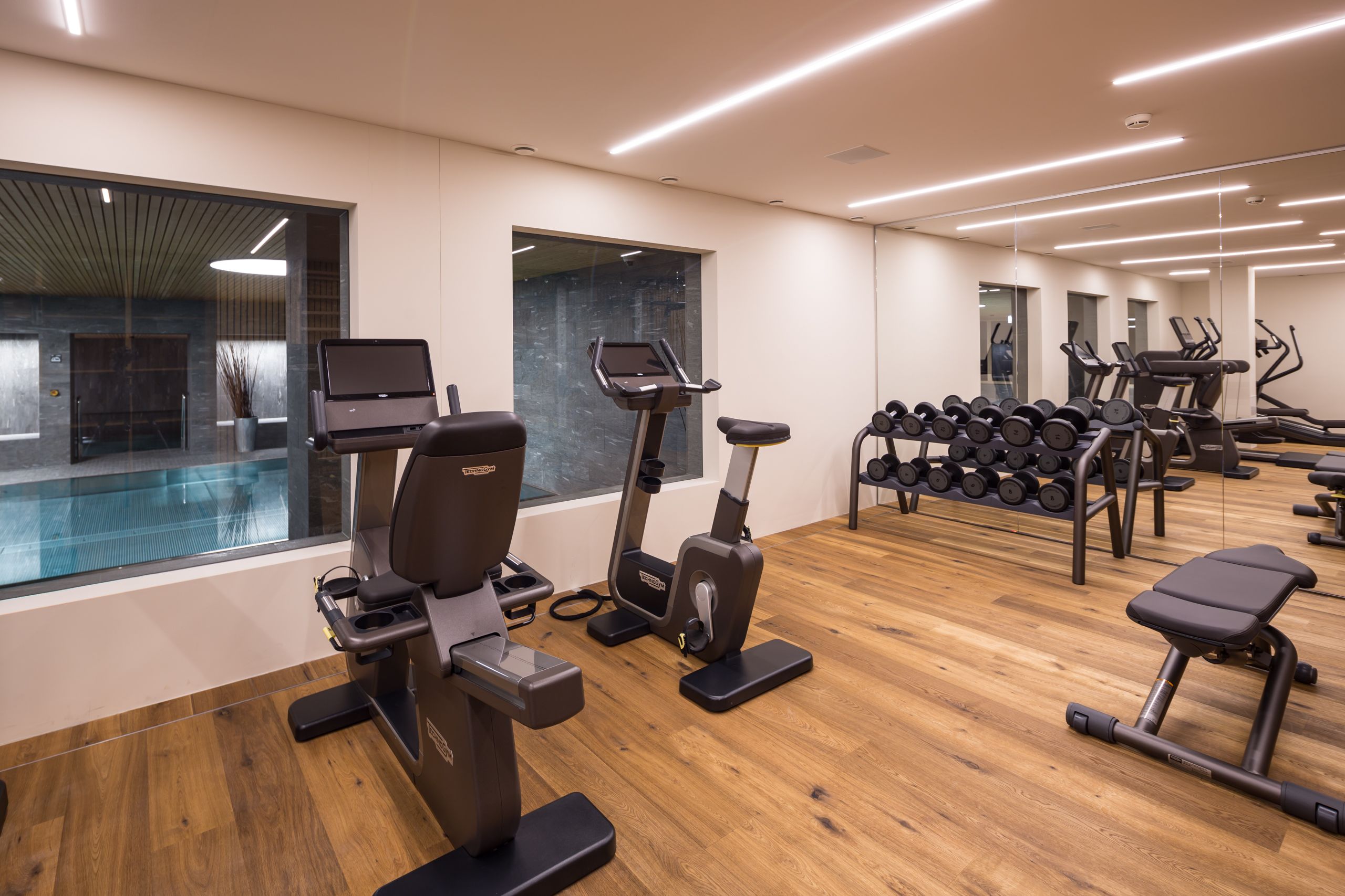 Ein modernes Fitnessstudio mit Laufbändern, Gewichtecken und einem Schwimmbecken, das durch die Glaswand sichtbar ist.