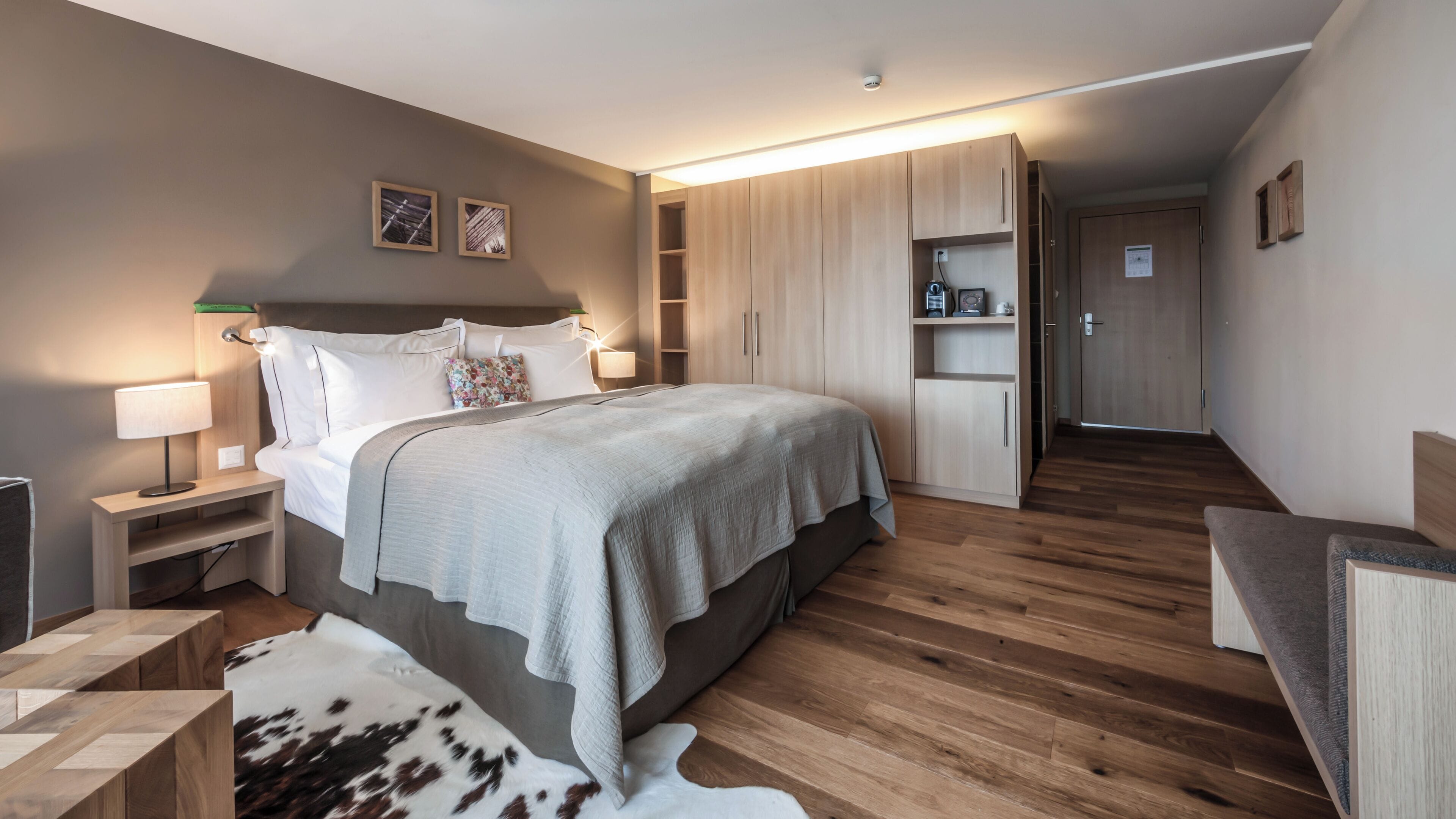 A modern bedroom with a large bed, bedside tables, a nightstand, a wooden wardrobe, and a small seating area.