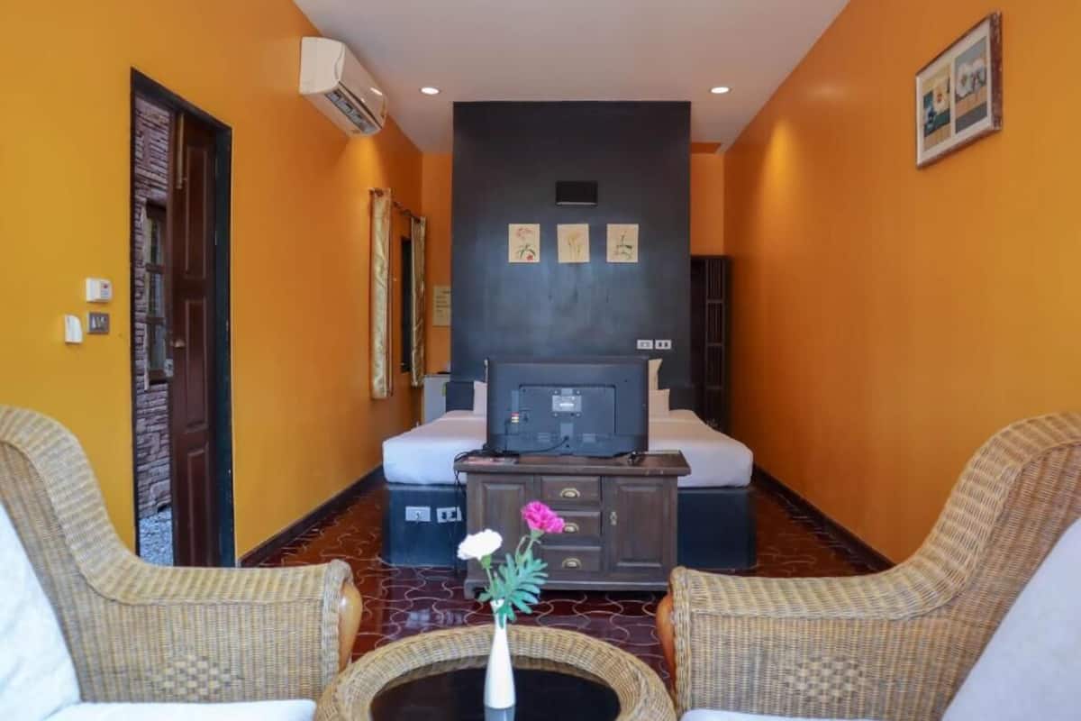 A hotel room with a bed, a television, a small table with a vase of flowers, two wicker chairs, and a dark-colored cabinet.