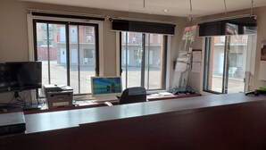 Een receptiebalie met een computer, printer en televisie. Grote ramen met zonwering en uitzicht op het exterieur.