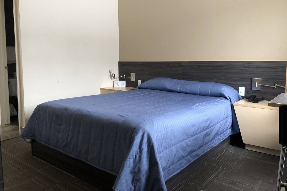 A neatly made bed with a blue bedspread, a bedside table with a lamp, and a wall-mounted light fixture.