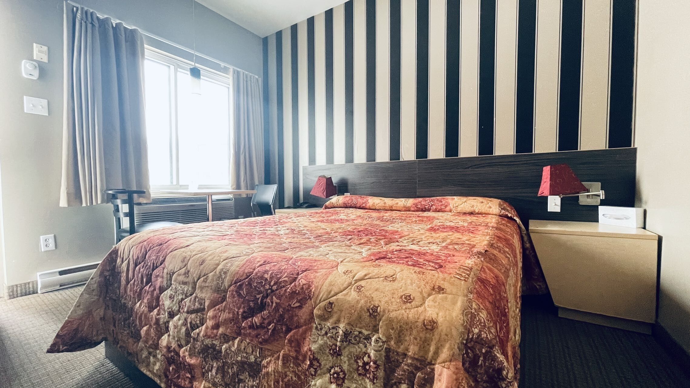 A hotel room with a bed, a bedside table, a lamp, a chair, and a window with curtains.