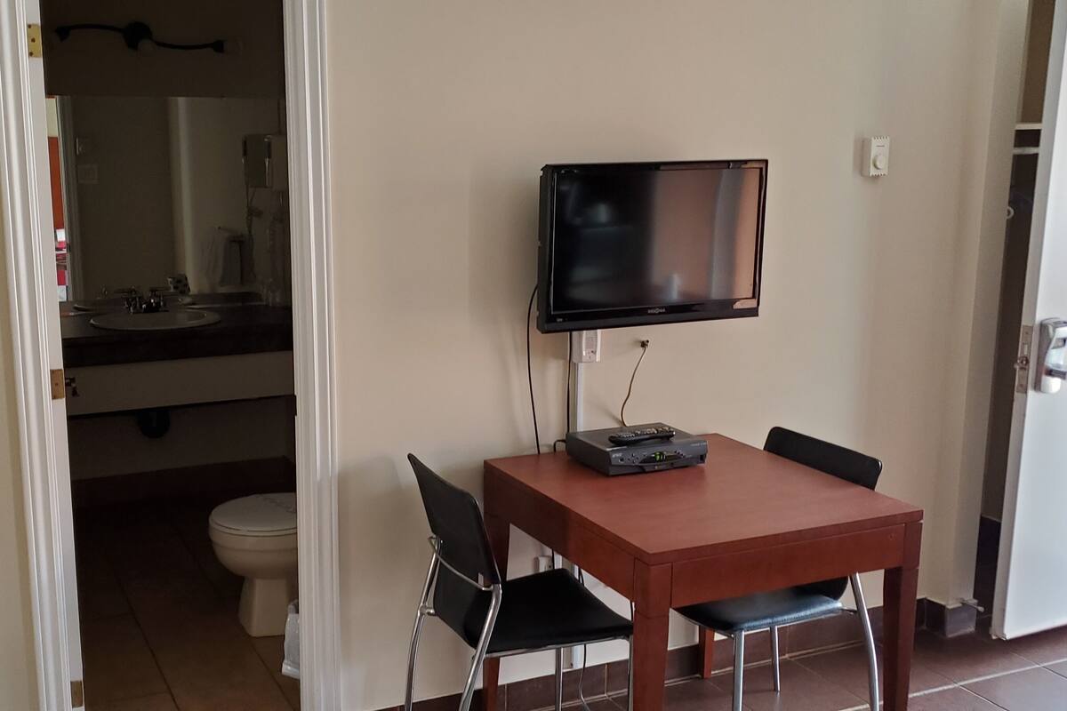 A small room with a wooden table, two chairs, a television mounted on the wall, and a bathroom visible through an open door.