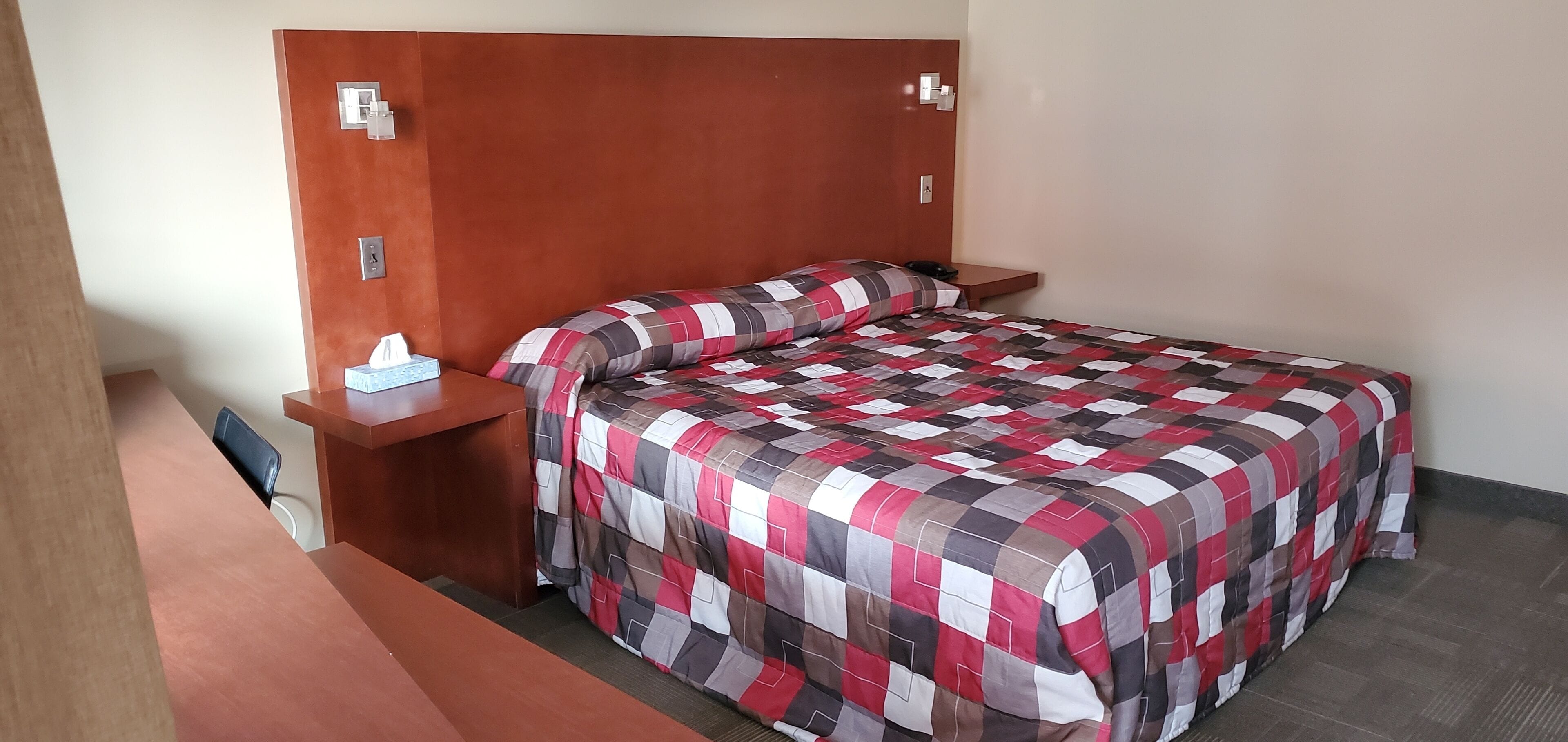 A neatly made bed with a checkered bedspread, a bedside table with a tissue box, and a wooden headboard.