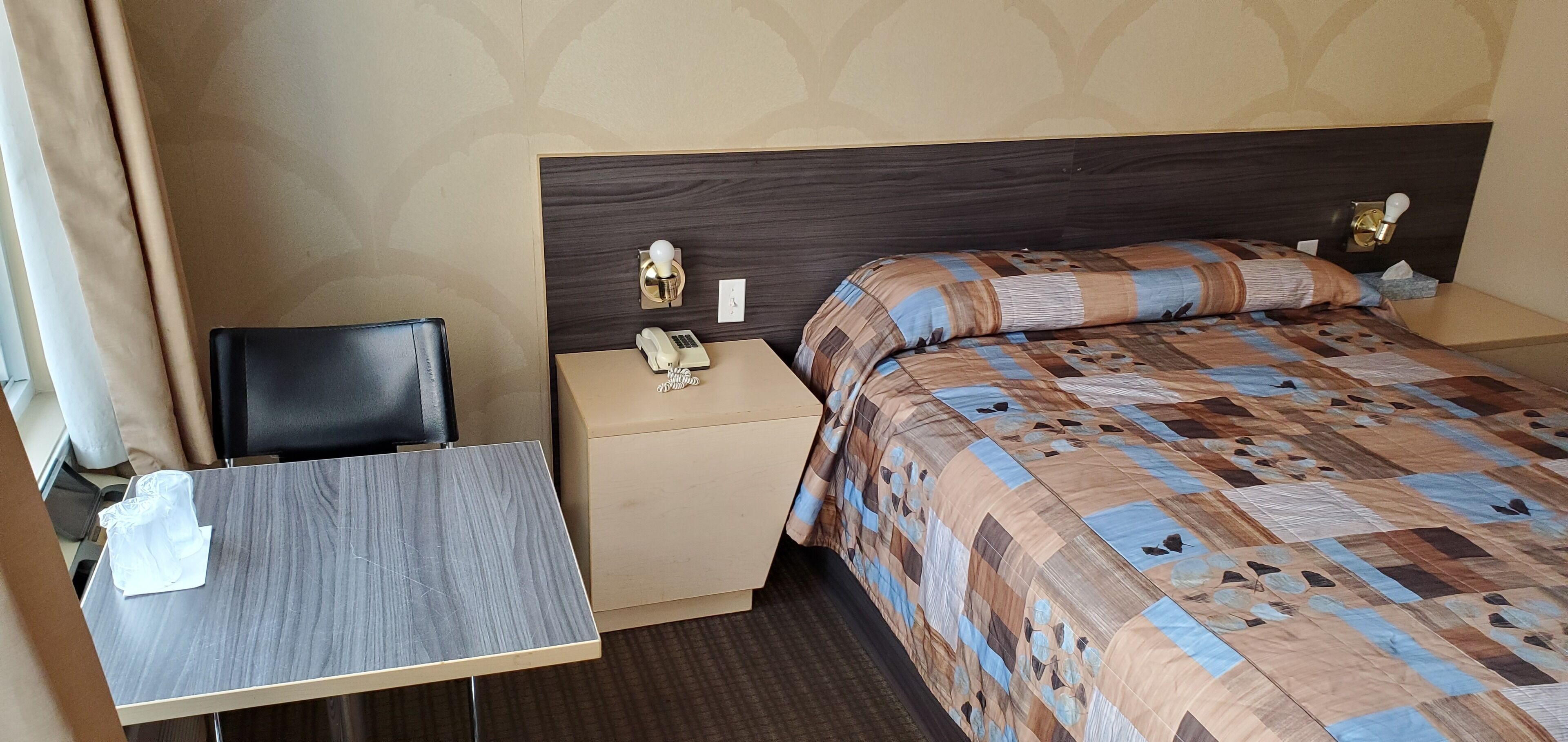 Habitación de hotel con una cama, una mesita de noche, una silla, un teléfono y una lámpara fijada a la pared.