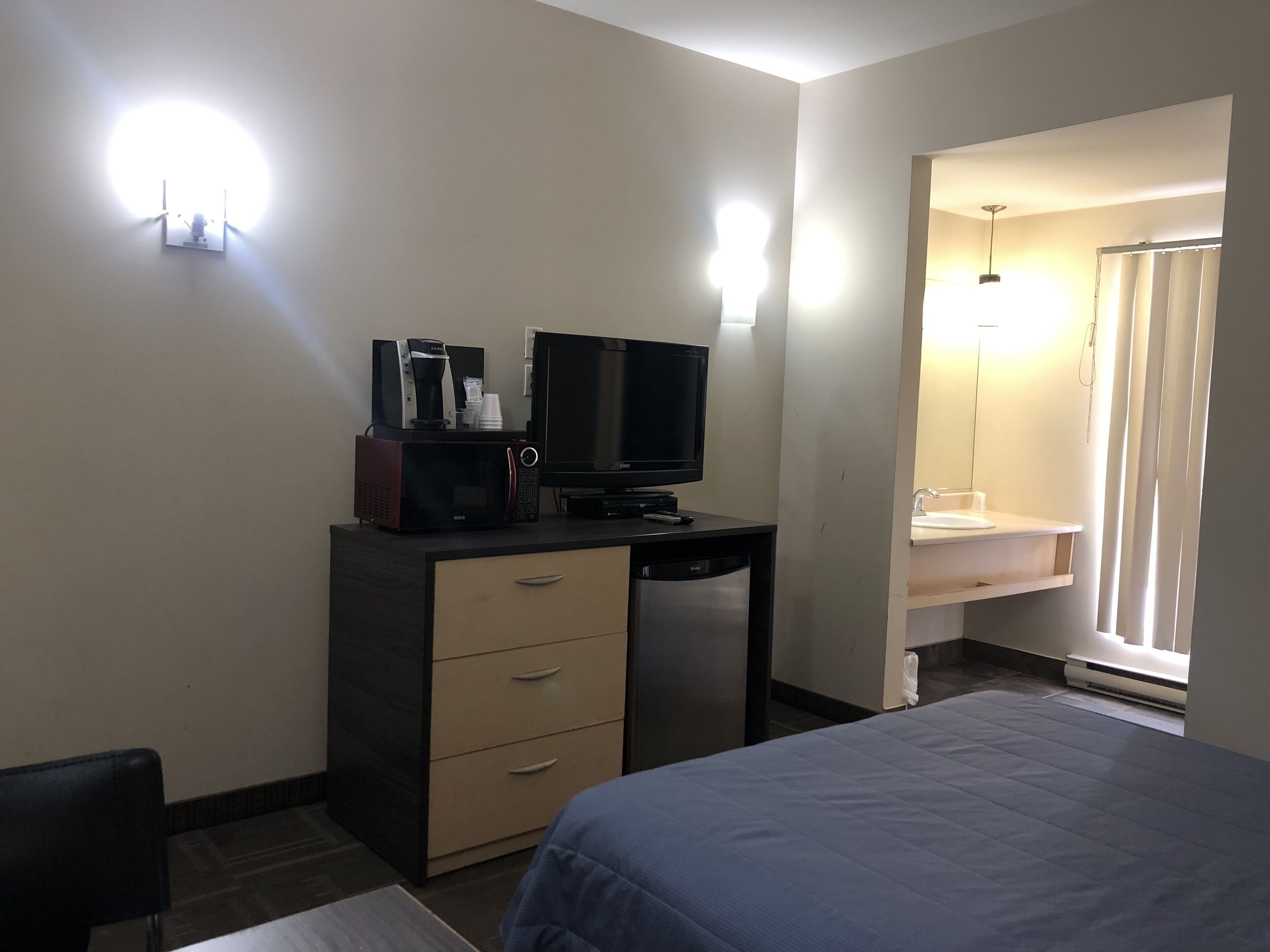 A hotel room with a bed, a desk with a television, a microwave, and a coffee maker. There is a bathroom with a sink and a shower.