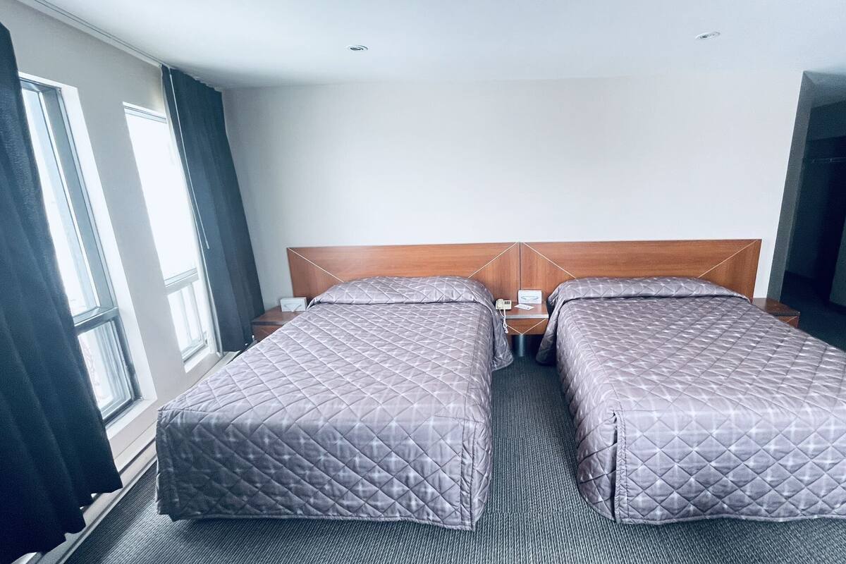 A hotel room with two single beds, a wooden headboard, a nightstand with a phone and a lamp, and a window with curtains.