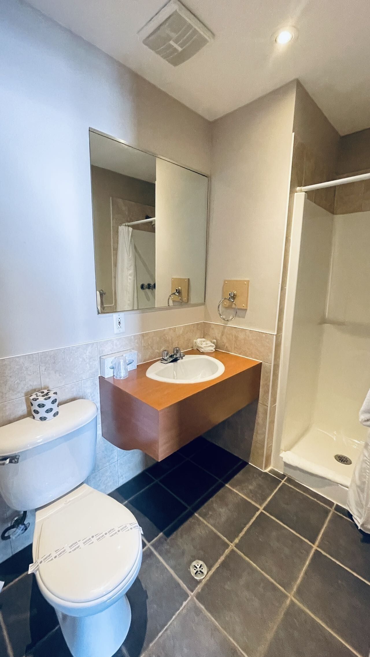Un baño con inodoro blanco, lavamanos con encimera color crema y espejo sobre el lavamanos, además de una zona de ducha.