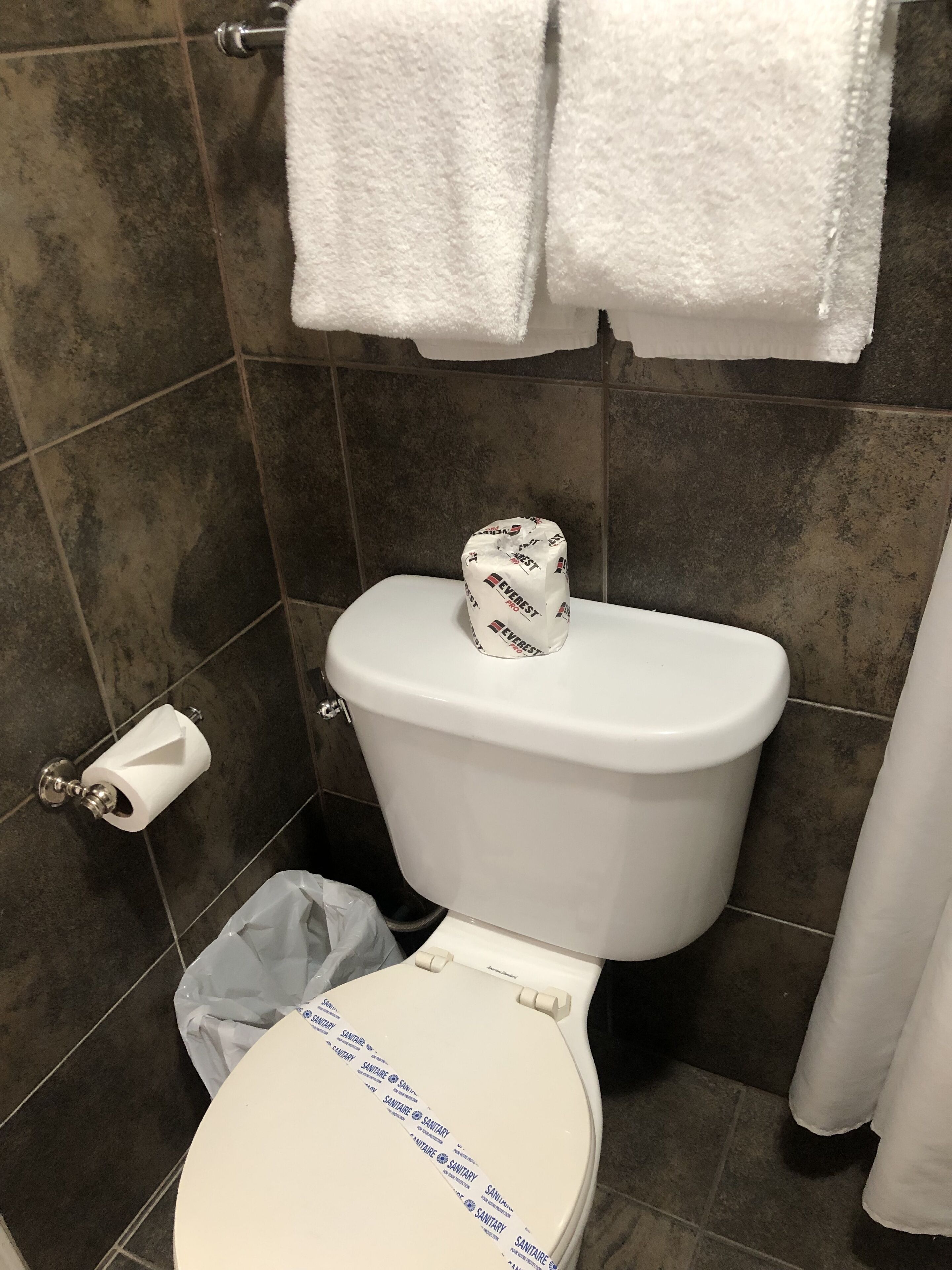 Un baño con un inodoro blanco, un rollo de papel higiénico, un cubo de basura y una toalla colgada en un perchero.