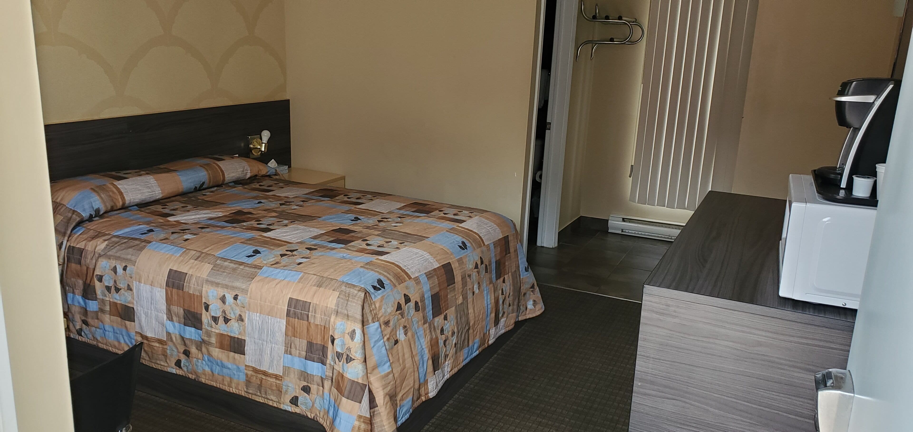 A neatly made bed with a patterned bedspread, a bedside table with a lamp, and a window with blinds.